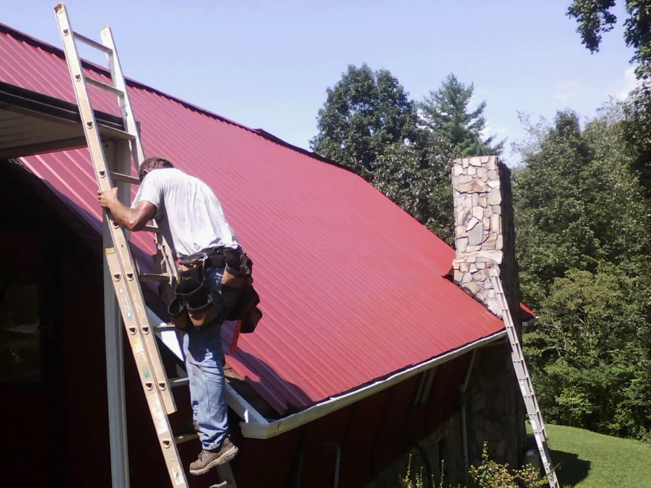 Roofer on a ladder beside a red metal roof and stone chimney — Soffit & Fascia Repair by Roofing Brothers in Skyforest